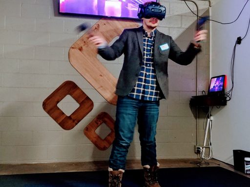 Andrew stands in front of a large tv showing laser lines. Andrew is wearing a VR headset and gesturing with controllers in both hands.