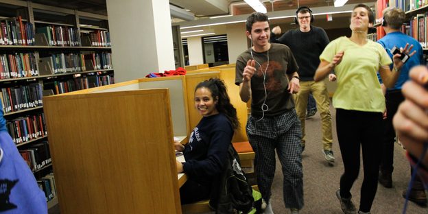 Students wearing earbuds dance through the library while another studies