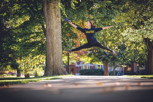 A student catches a frisbee while leaping across a campus path