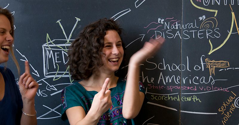 Two students gesture and laugh in front of a chalkboard.