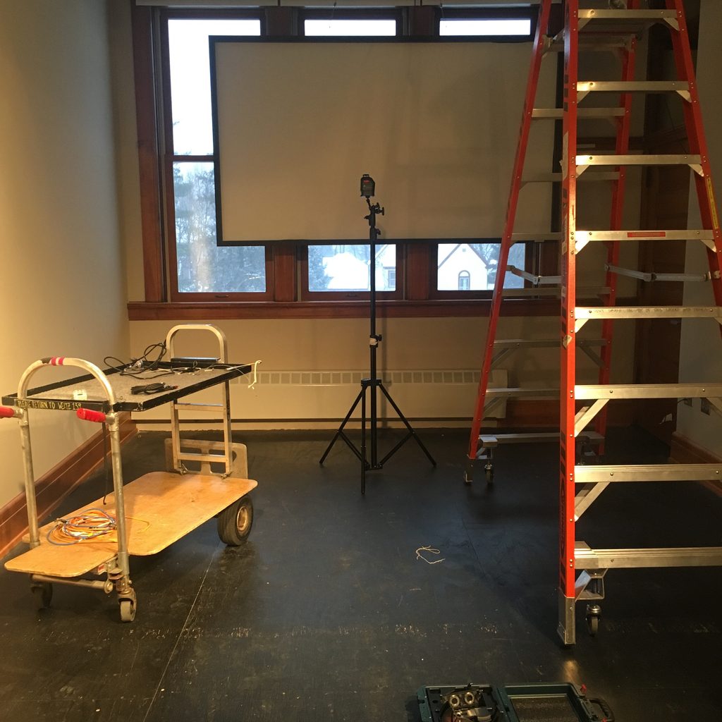 a small white room with a projection screen, a ladder, a small cart, and a black metal stand in the center