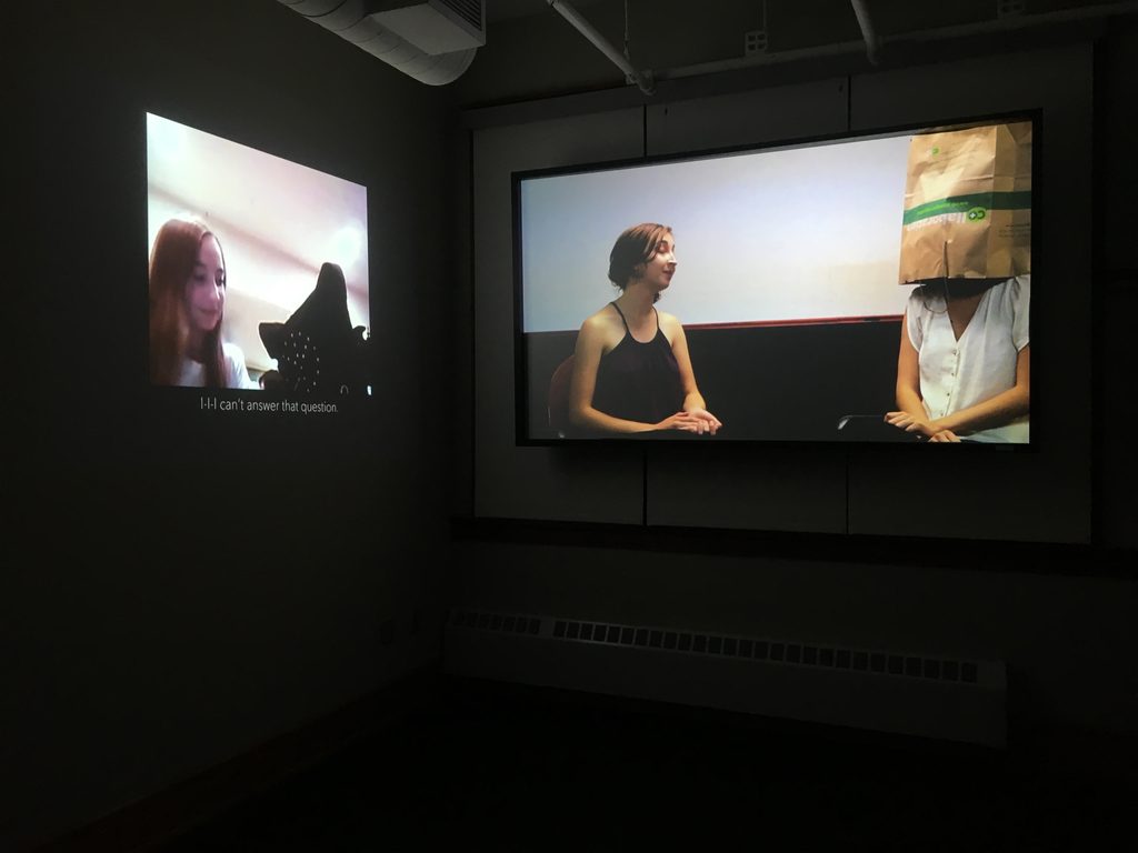 Two projected images at a 90 degree angle, showing a girl sitting next to a person with a bag on her head
