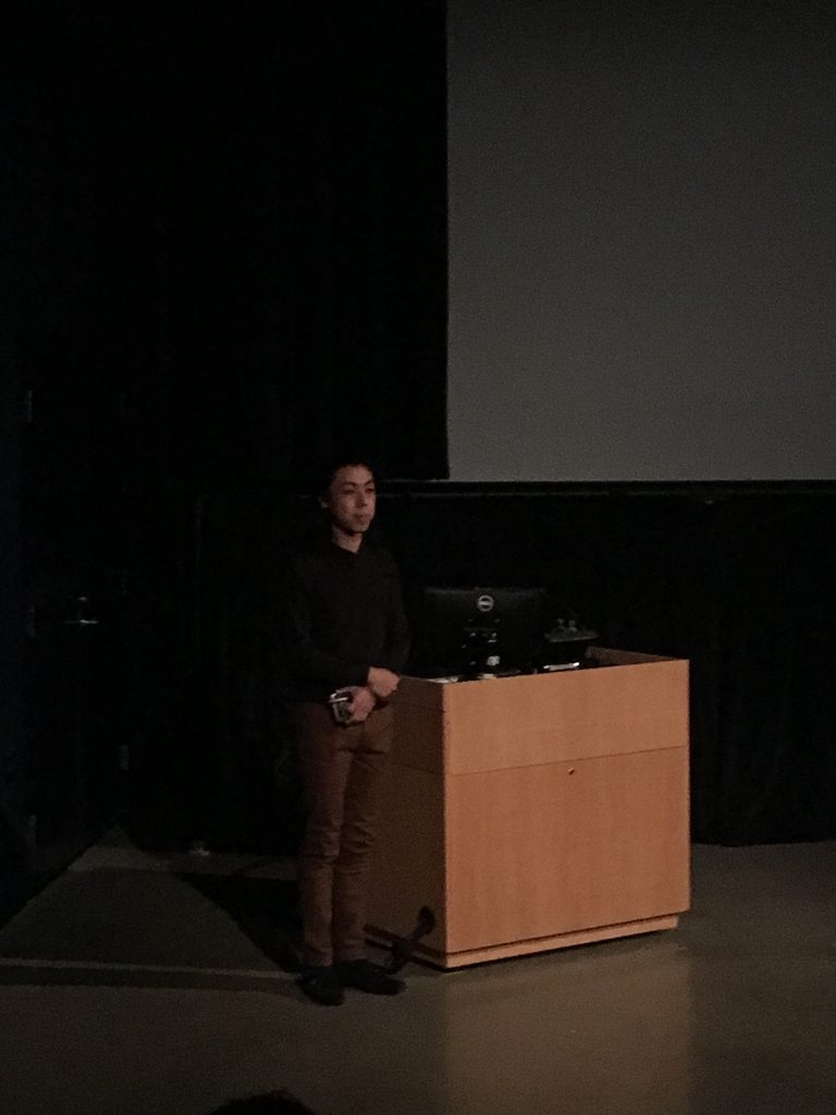 man standing beside podium in front of a projection screen in a theater