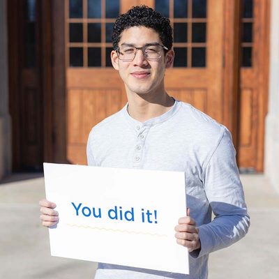 A student smiling and holding a sign that reads "You did it!"