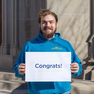 A student smiling and holding a sign that reads "Congrats!"