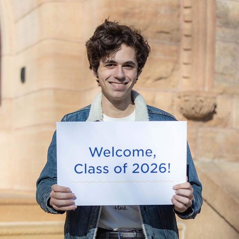 A student smiling and holding a sign that reads "Welcome, Class of 2026!"