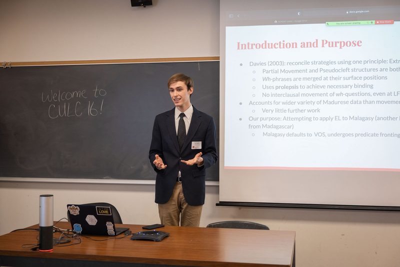 Tate stands in front of blackboard and screen, presenting his talk "Extreme Locality Applied Globally: The Case for Extreme Locality in Malagasy."