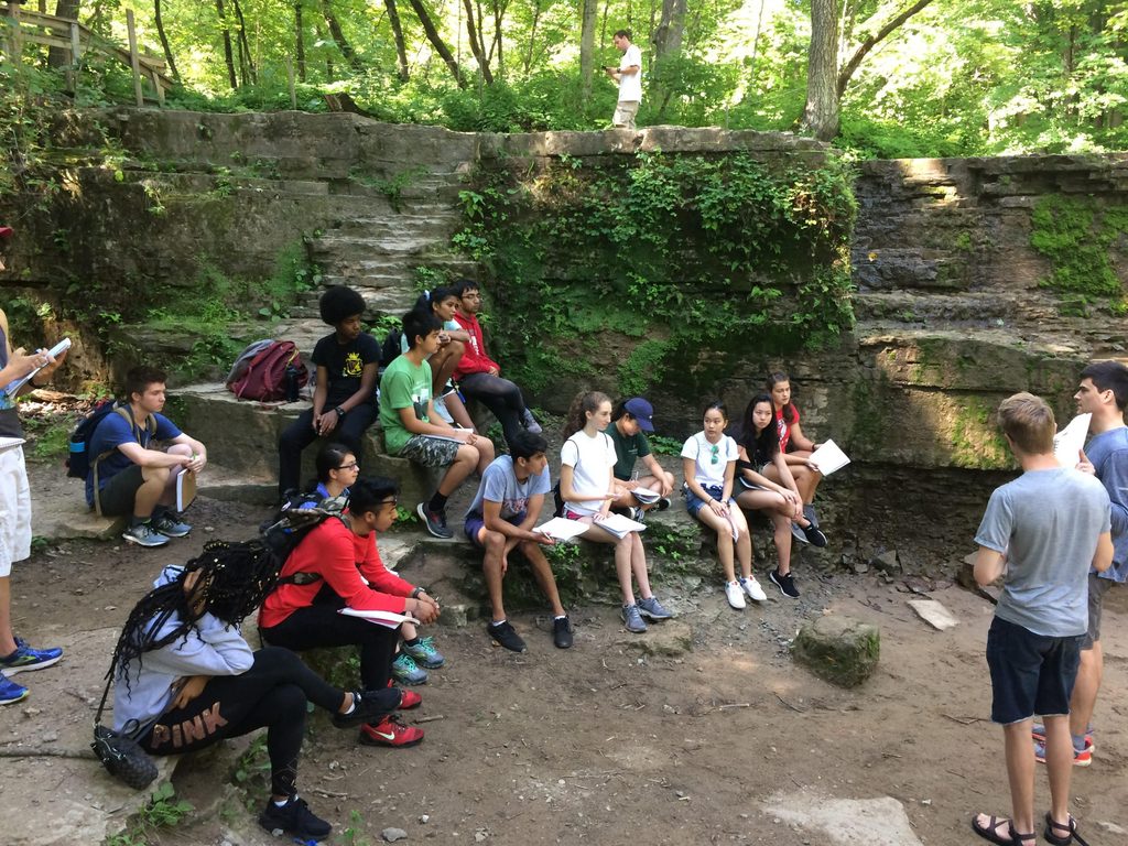A group of about 15 students sits in a forested area taking notes and facing the professor
