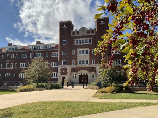 The entrance to Burton Hall, a red brick building, sprawls the background to this photo with some tree branches covered in pink berries coming into the right foreground.