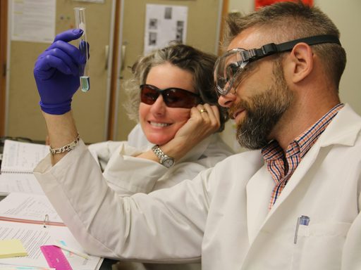 An APSI teacher wearing a white lab coat, safety goggles, and a purple latex gloves holds up and looks at a test tube with a bit of blue liquid in it while another teacher leans in to look at the test tube also.