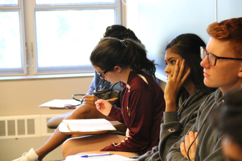 Students in a classroom during lecture. Two are looking at the professor and one's head is bent down writing notes.