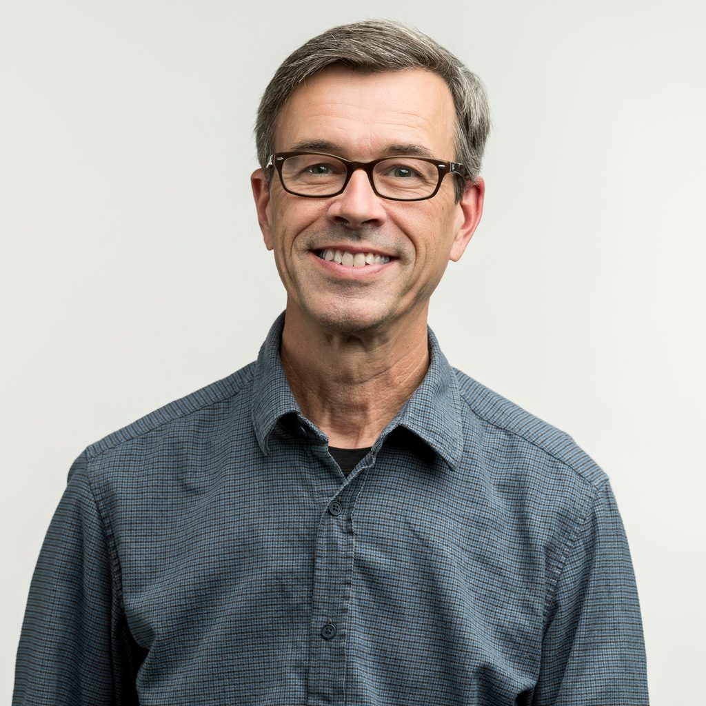 Headshot of professor Steve Drew from the chest up. He is wearing a blue button up, glasses, and is smiling in front of a white background.