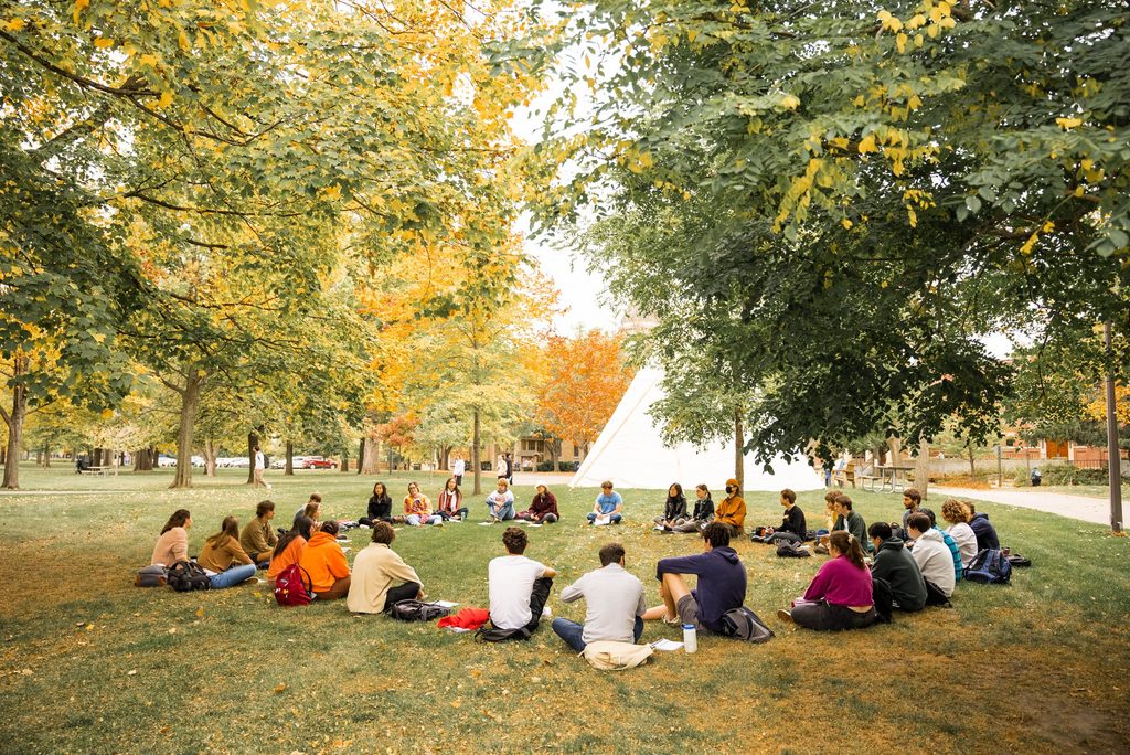Carleton students sit in a circle outside on Carleton's bald spot. The ground is grassy and trees shade the students. A white tipi is set up in the background.