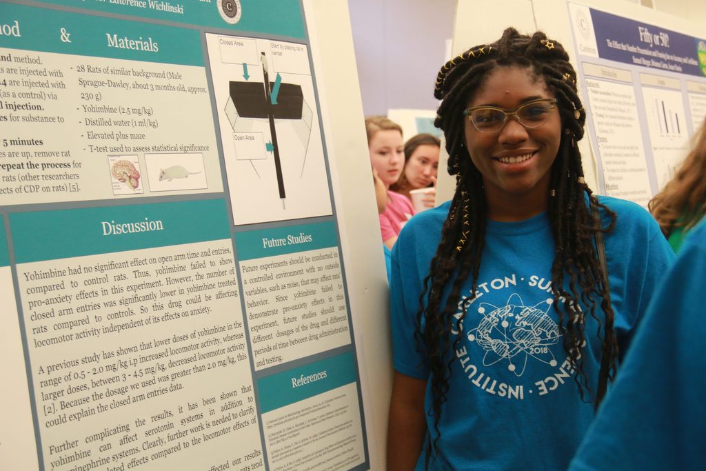 Student smiling next to their neuroscience research poster. The student is wearing a blue shirt with a drawing of a brain on the front.
