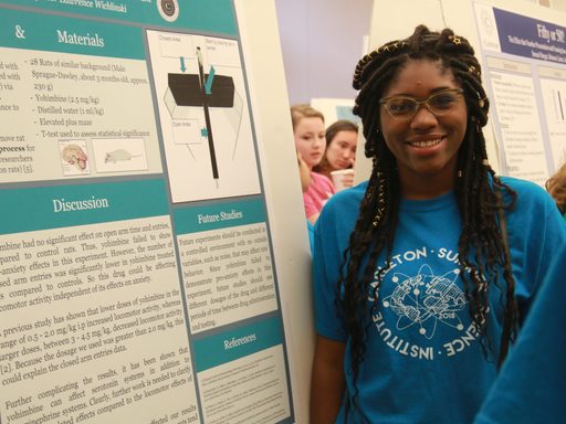 Student smiling next to their neuroscience research poster. The student is wearing a blue shirt with a drawing of a brain on the front.