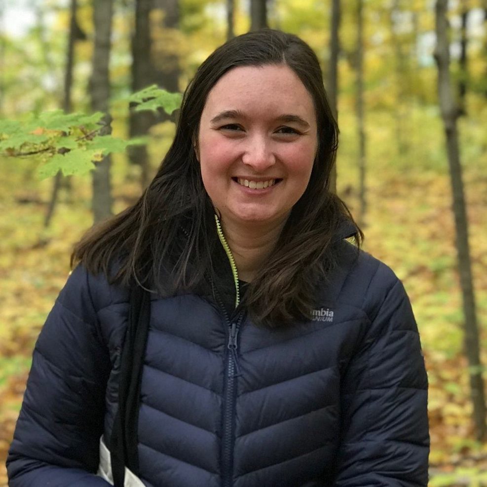 Professor Laura Biester smiling in a forest. Laura has long dark hair and wears a black puffy jacket.