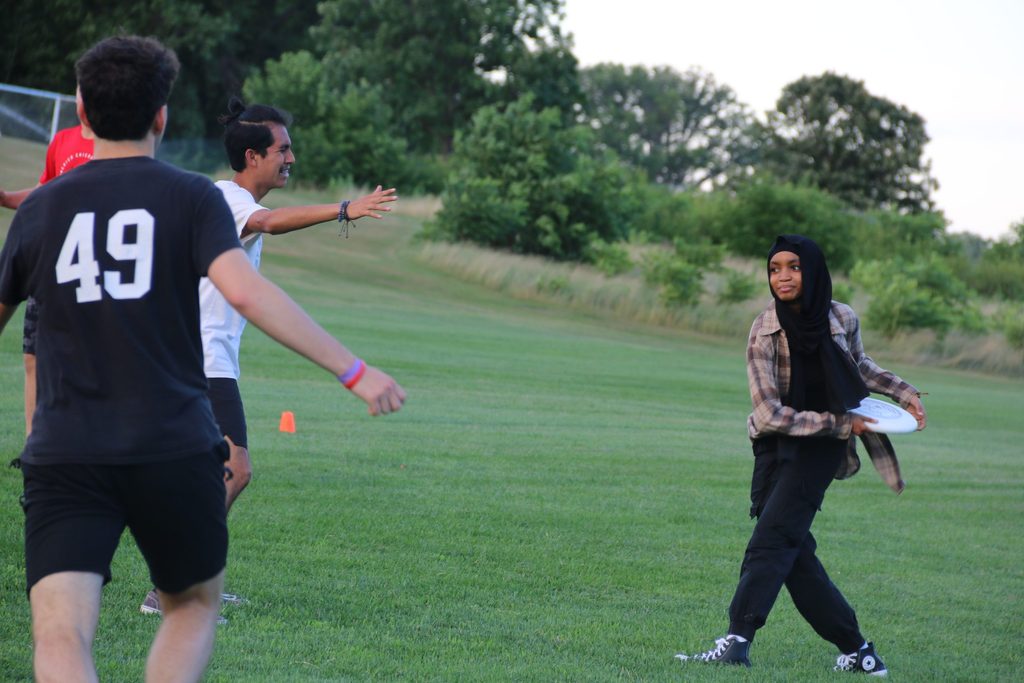 Pre-college students playing frisbee on a green field: One student on the right of the photo holds the frisbee and is preparing to throw while two students on the left of the photo hold their arms out in defense.
