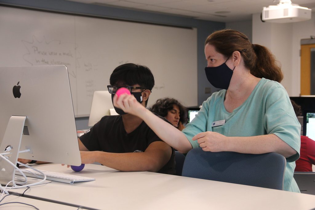 Professor Susan Fox assisting a pre-college student. She holds up a small pink ball while looking at a computer screen with the student to her right.