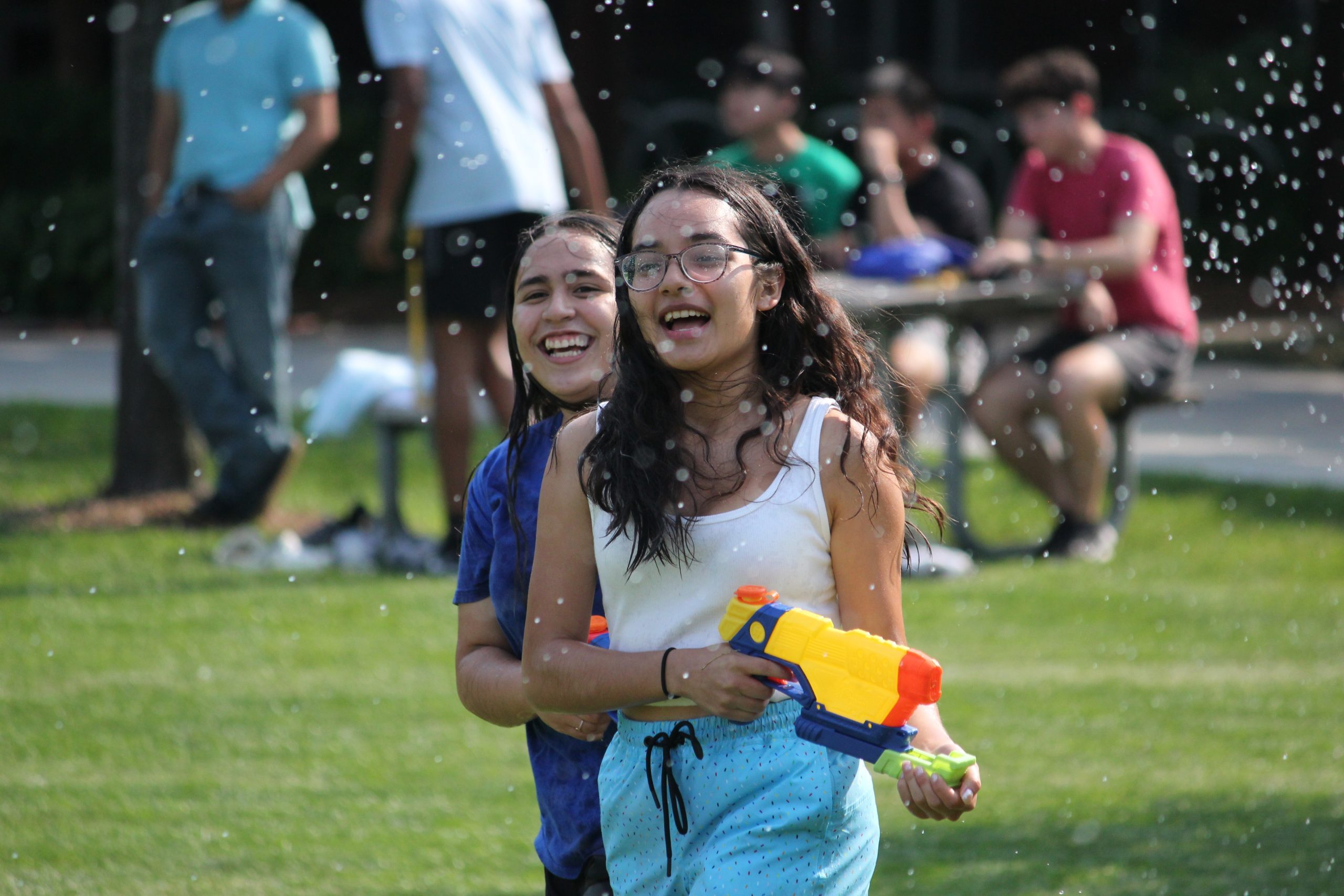 Waist up photo of two pre-college students running and laughing on the grass. The student in front is holding a water gun and water droplets are spraying on the students.