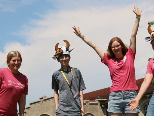 A Summer Carl stands and smiles wearing a cow hat and their Carleton lanyard, surrounded by Carleton summer student staff members posing excitedly.