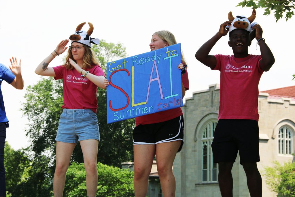 Three people dancing and smiling with the Carleton chapel in the background. The outer two are wearing cow hats with their arms raised, and the midle person is holding a sign that says "Get Ready to SLAI"