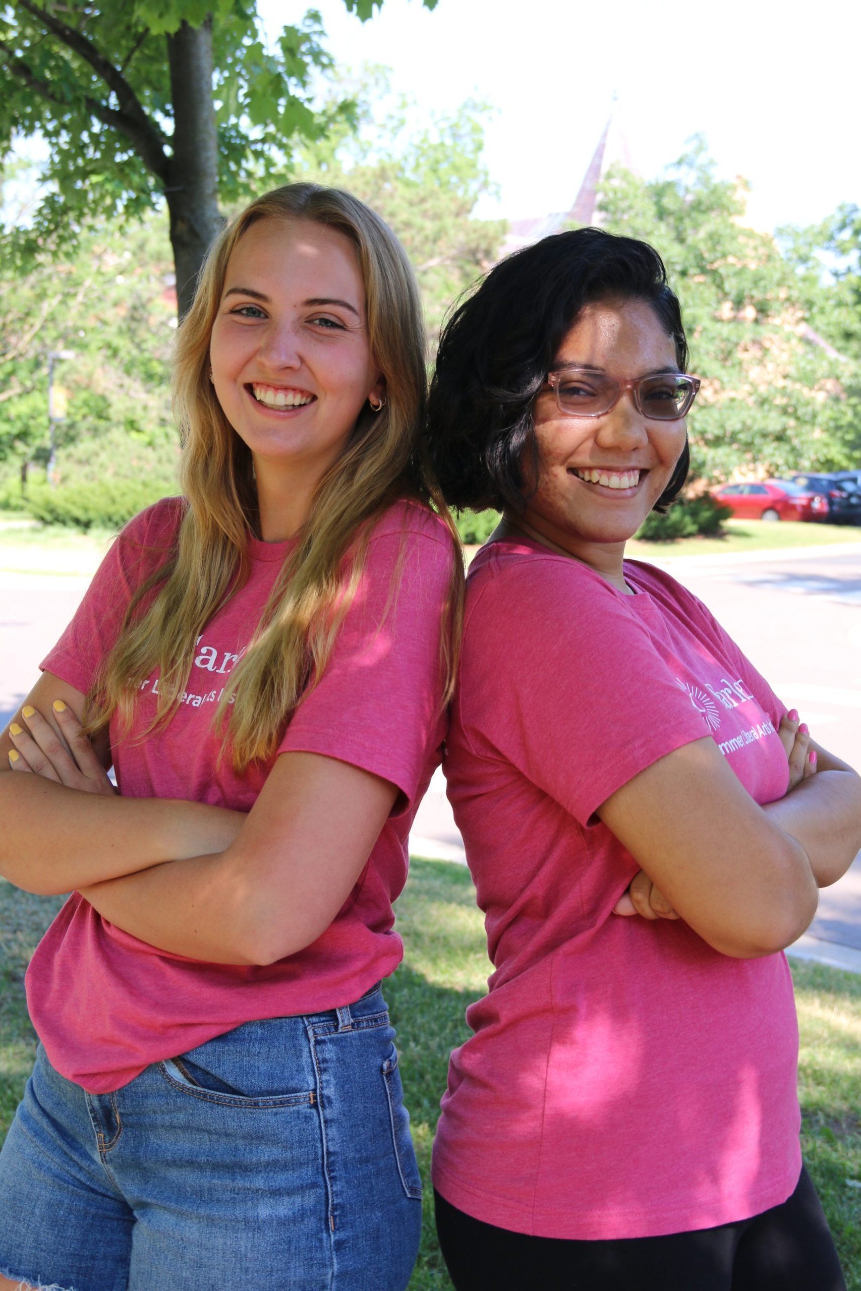 Two students standing back to back with their arms crossed, smiling and looking at the camera. They are wearing pink t-shirts that have the Carleton logo on the front.
