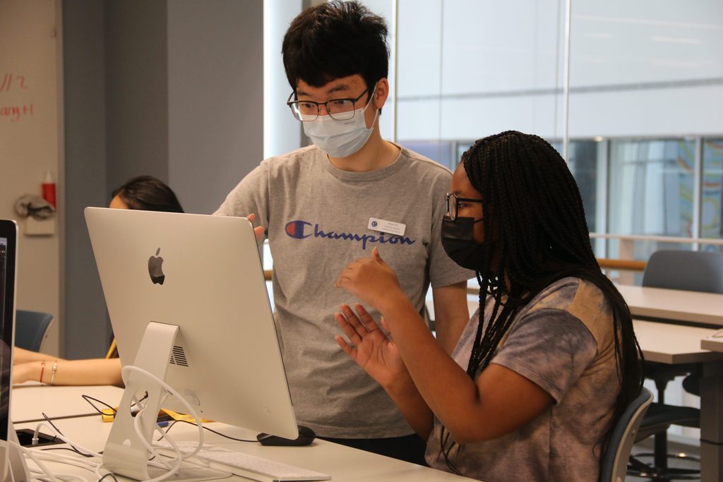 A teaching assistant and CS student look at a computer. The student is sitting and gesturing with her hands while the TA stands, looking intently at the screen.