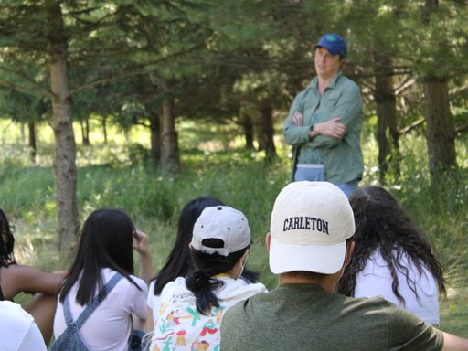 A group of students seated on the grass watching a professor lecture outside. We can see the backs of the students heads and the professor is blurred in the background. One student wears a Carleton hat backwards