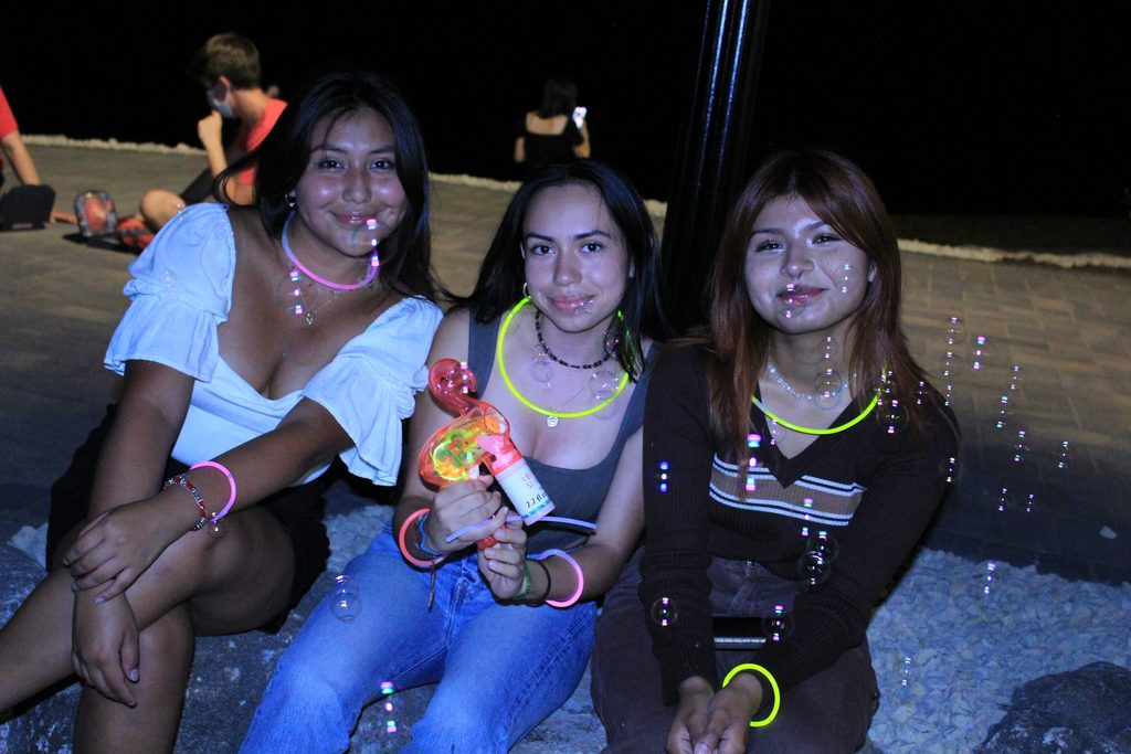 Three students sit outside in the evening. They are wearing glow necklaces and bracelets and shooting bubbles out of a bubble gun. They smile at the camera