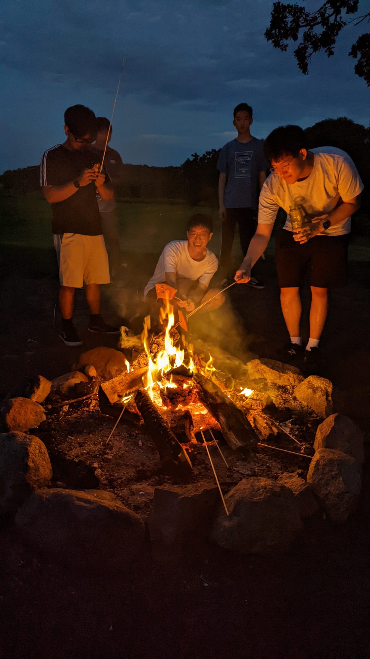 Two students crouch down by a campfire at night. They are holding sticks and roasting marshmallows over the fire.