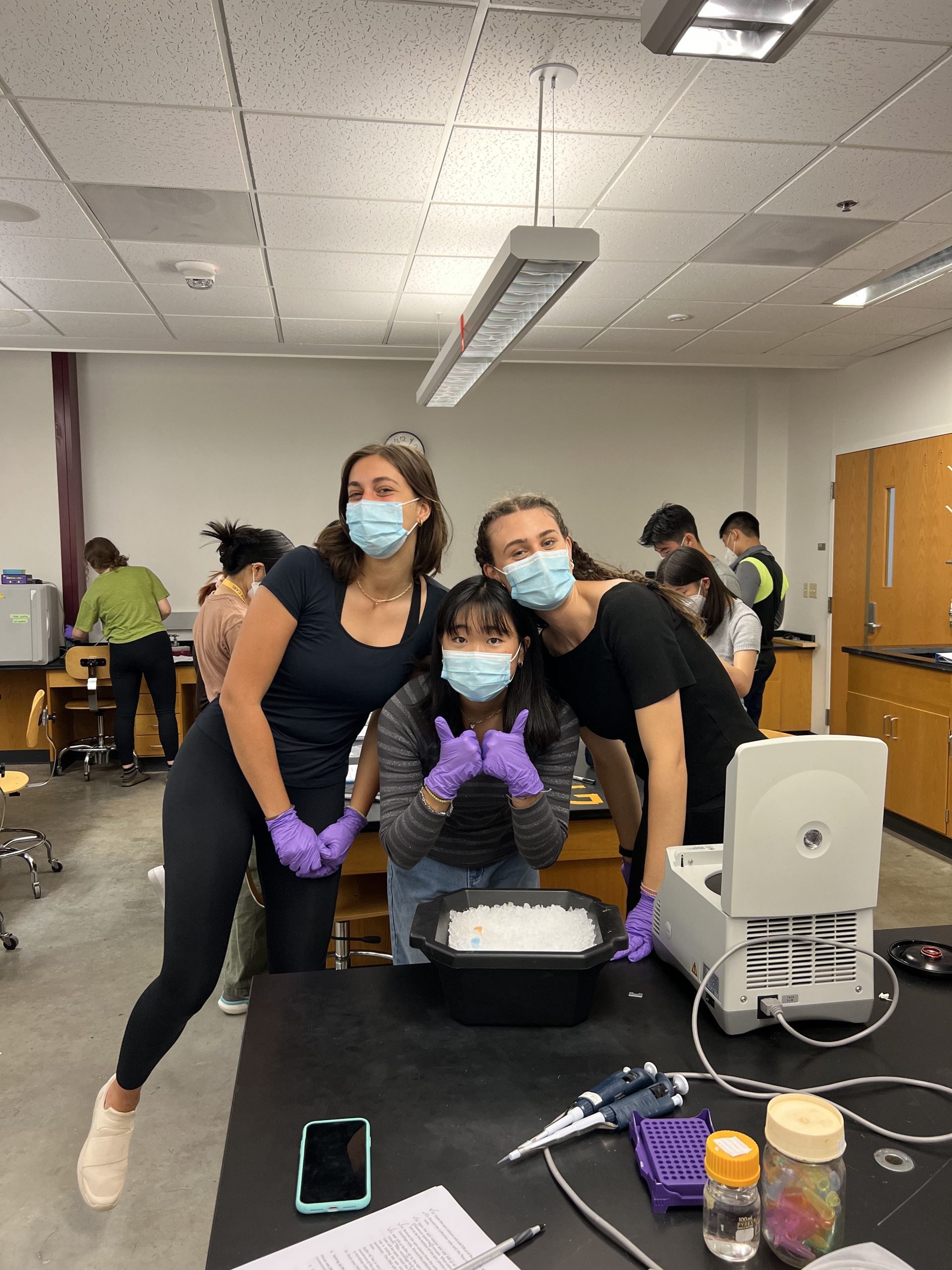 Three students smile by a biology lab instrument. They wear masks and purple gloves, and the middle student is giving two thumbs up