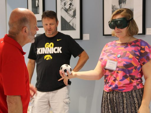 An APSI participant stands and holds a small soccer ball in front of her body with her right hand. She is wearing opaque goggles, and the teacher stands facing her giving instructions