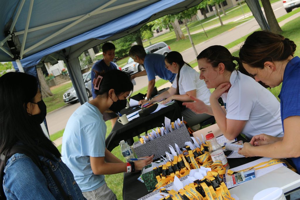 Two SLAI staff sit behind a table with boxes of keys and lanyards. They talk to two students standing on the other side of the table, and one is signing a piece of paper
