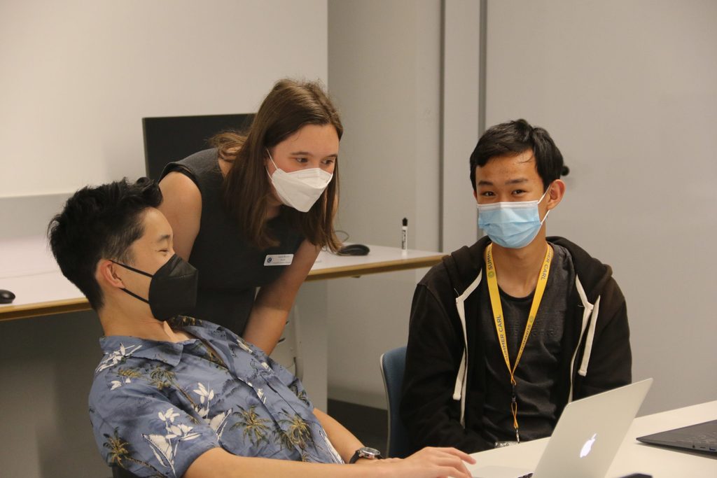 A computer science professor helps two students on their laptop.