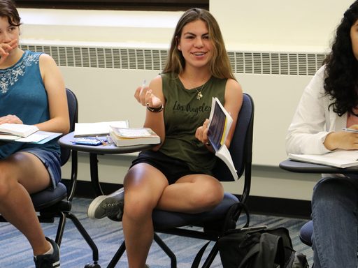 A student sits at her desk and gestures with a book in her hand while looking beyond the camera, presumably at the professor.