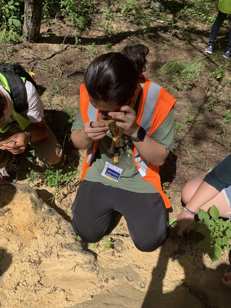 A STEM student kneels on the ground using a hand lens to look at a rock sample