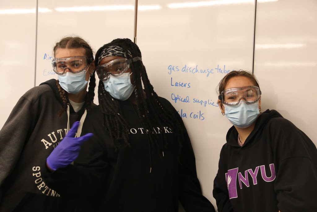 Three STEM students smile wearing goggles in front of a white board