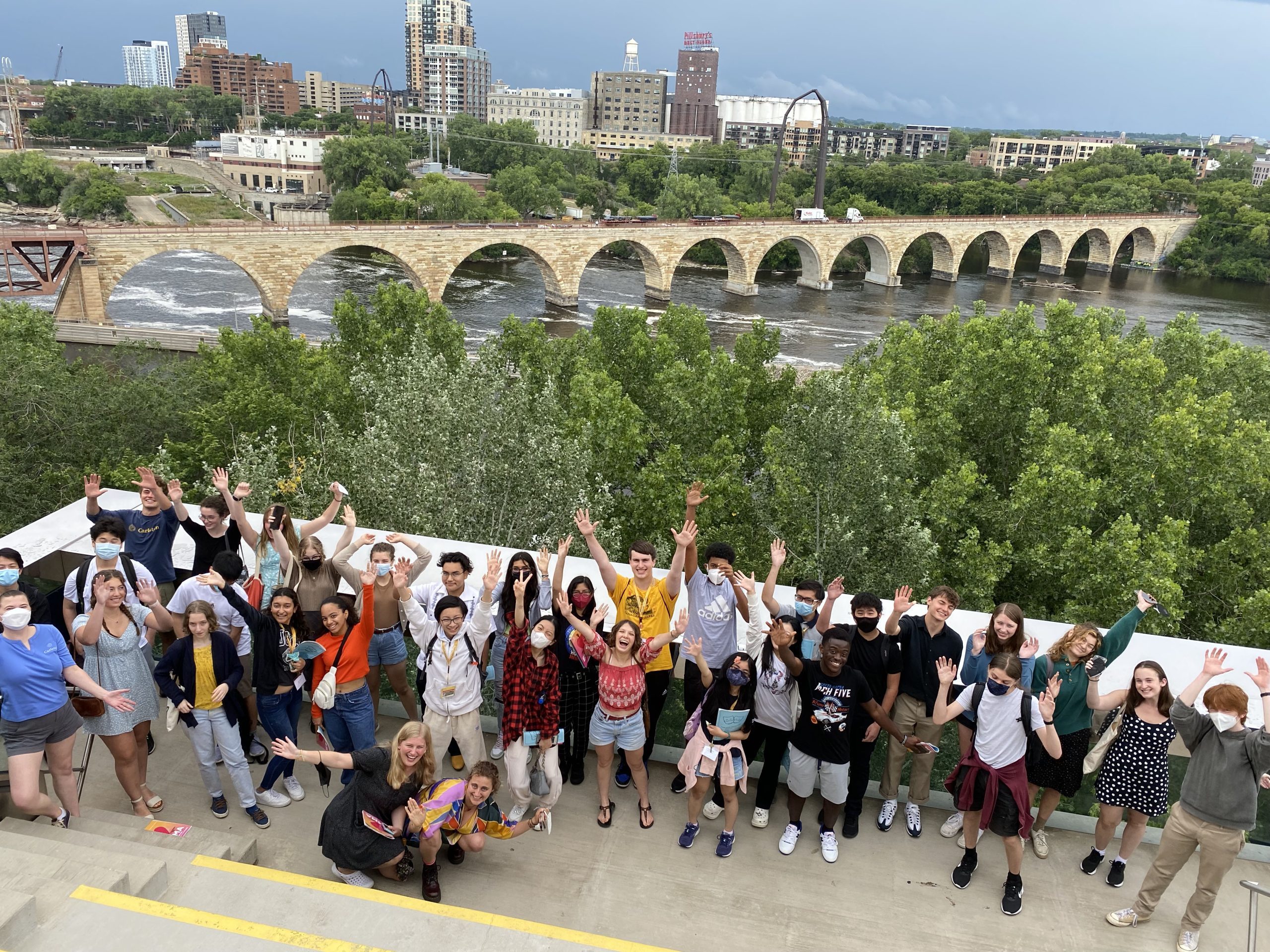 A group of students gathered on a balcony overlooking the Minneapolis skyline and Stone Arch Bridge. They look up at the camera, smiling with their hands in the air