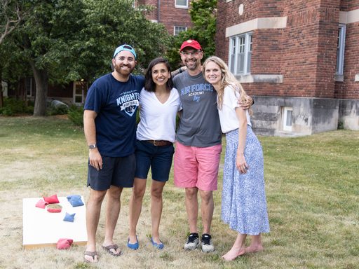 4 adults in summer garb stand on the grass outside a Carleton dorm and smile with their arms around each other. There is bean bag toss behind them.