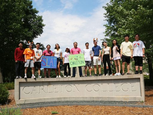 13 students standing atop the Carleton College sign and smiling. Some are wearing cow hats while others hold signs that read 