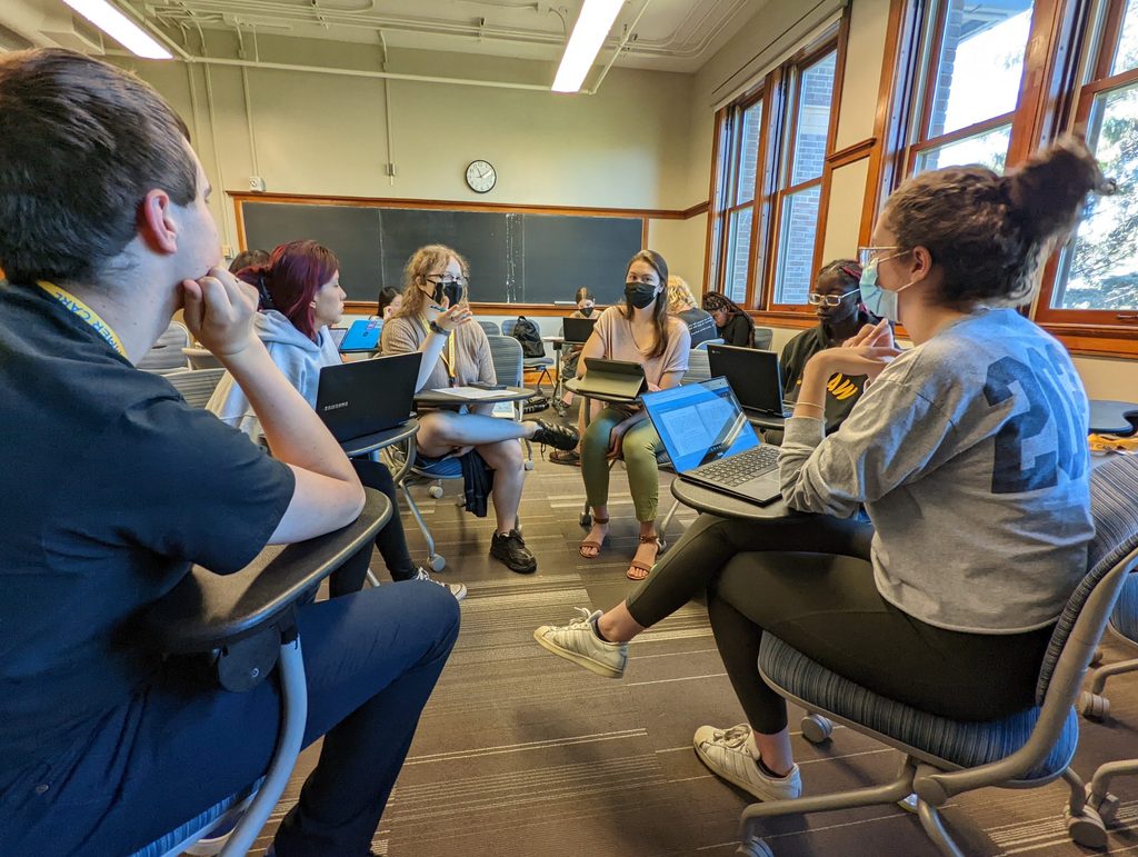 Humanities students sit in a circle of desks with laptops in discussion