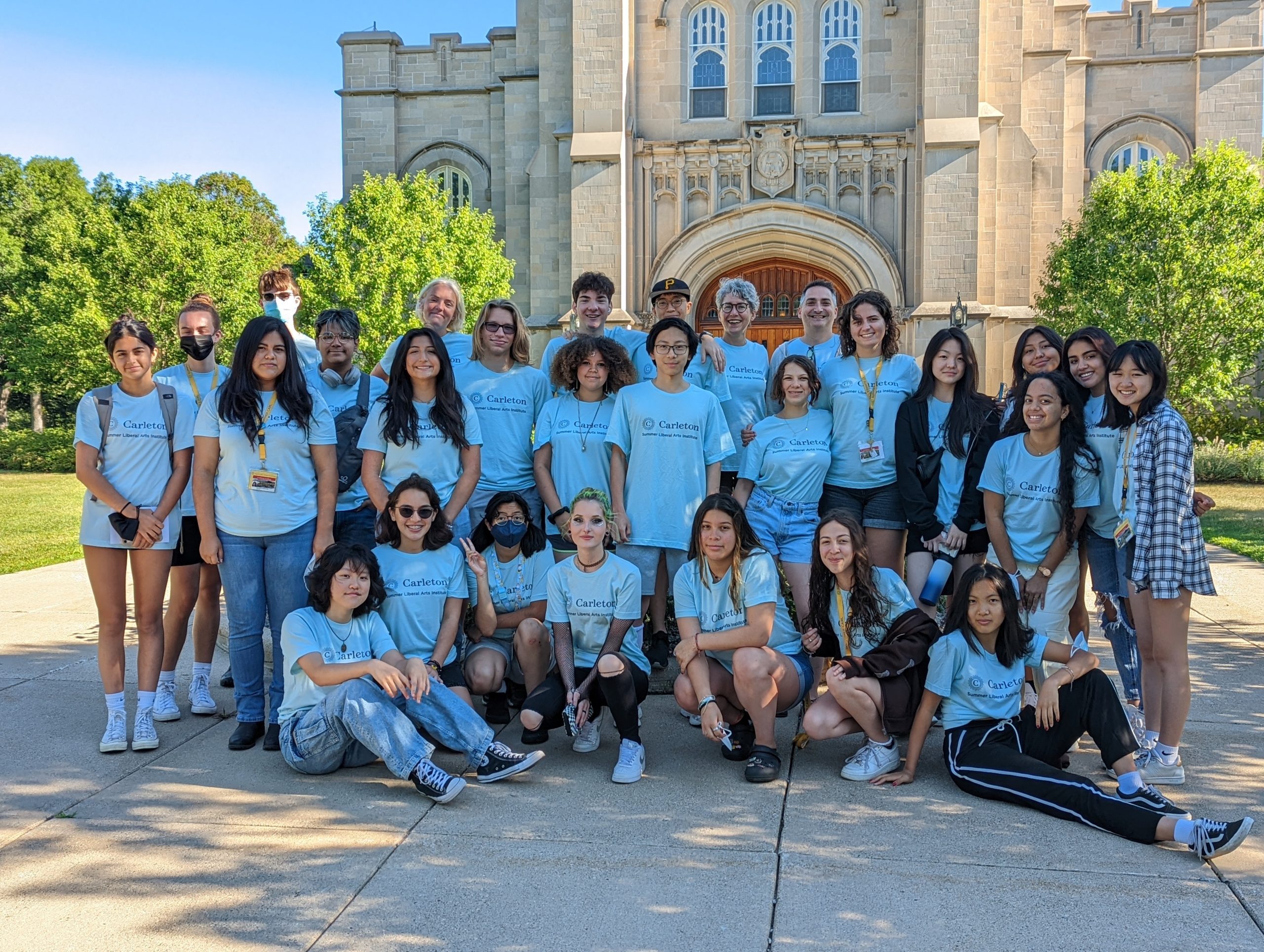 A group of 24 students and their professors gather and smile in front of Carleton's Chapel. They are all wearing light blue Carleton SLAI t-shirts