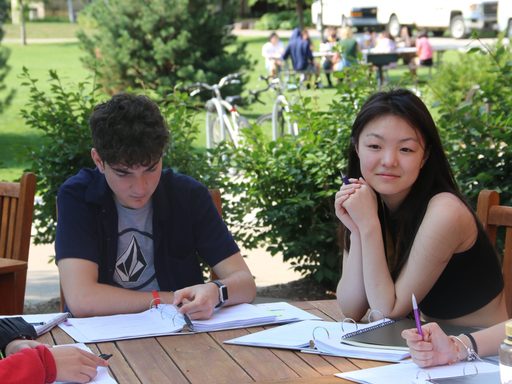 Two students sit at an outdoor picnic table with their material binders open in front of them. One looks down at his binder while the other props her chin on her hands and looks towards another student with a slight smile