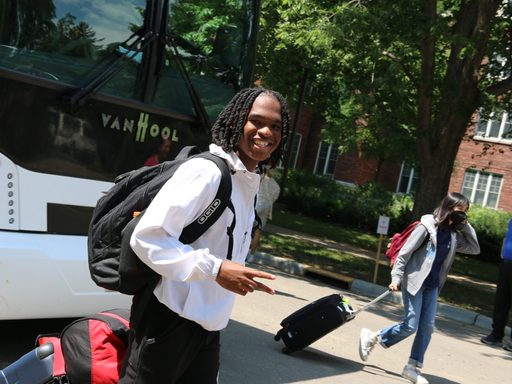 A walks in front of a bus with his suitcase and smiles with throwing up a peace sign at the camera