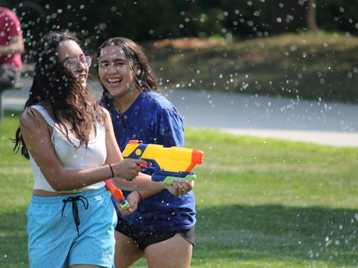 Two students laughing as they walk through a spray of water. Both hold water guns and one is shooting it off to the right