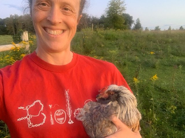 Professor Annie Bosacker smiling in front of a field, wearing a red Biology shirt, and holding a chicken.