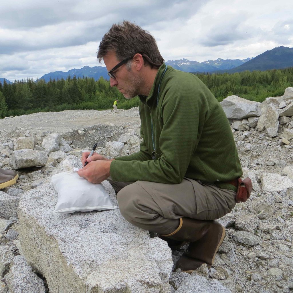 Professor Cam Davidson crouching over a big piece of granite, writing on a rock sample bag. The background consists of broken up rock, trees, and mountains.