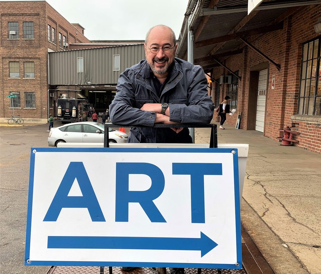 Professor Pierre Hecker stands behind a sign that says ART in big letters with a right arrow. He leans his forearms on the sign and smiles