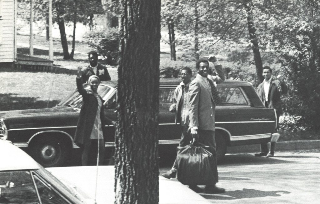 A group of students stand near a car in the 1960s.