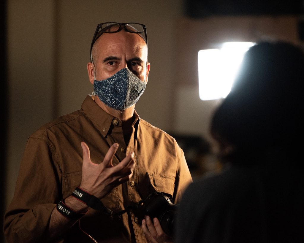 A male professor talks while wearing a mask, with a light visible in the background. 
